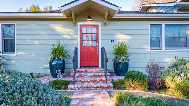A house with a red door, two potted plants on either side, and a brick pathway leading to the entrance.