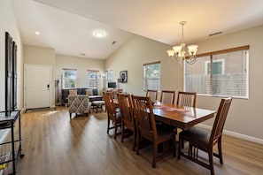 A dining area with a wooden table and chairs, a chandelier, and a view of the outdoors through large windows.