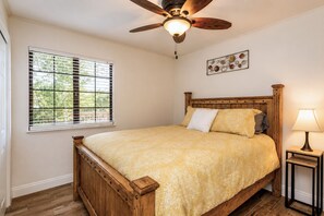 A bedroom with a wooden bed, a ceiling fan, a window with blinds, and a bedside table with a lamp.