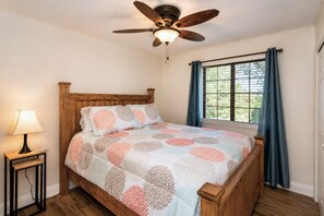 Un dormitorio con cama de madera, ventilador de techo, lámpara de noche y ventana con cortinas.