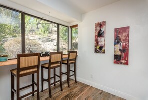 Comedor con mesa y sillas de madera, ventanas amplias con vista a árboles y dos cuadros en la pared.