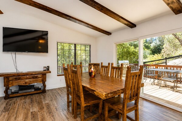 A dining area with a wooden table and chairs, a television mounted on the wall, and a view of the outdoors through large windows.