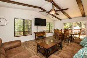 A spacious living room with a wooden ceiling, a ceiling fan, a flat-screen TV, a dining area with wooden furniture, and large windows with blinds.