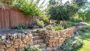 A stone wall with steps leading to a garden area, surrounded by various plants and a wooden fence.