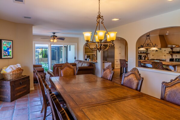 A spacious dining area with a long wooden table and ornate chairs, a chandelier, and a view into the kitchen.