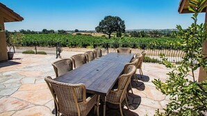 A patio with a table and chairs, a stone floor, a fence, and a vineyard in the background.