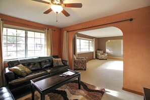 A living room with a dark leather sofa, a coffee table, and a ceiling fan. There are two windows with curtains, a patterned armchair, and a carpeted floor.