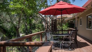 Eine Holzterrasse mit einem Metalltisch und -stühlen, einem roten Sonnenschirm und einem Geländer mit Blick auf einen Garten.