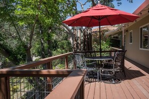 Eine Holzterrasse mit einem Metalltisch und -stühlen, einem roten Sonnenschirm und einem Geländer mit Blick auf einen Garten.
