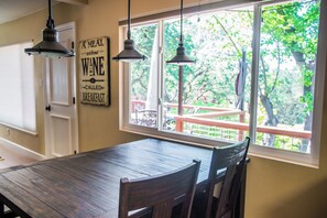 Un espace repas avec une table et des chaises en bois, une fenêtre donnant sur un paysage verdoyant et des luminaires suspendus.