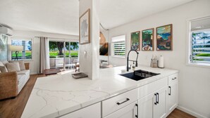 A modern kitchen with a large island, white cabinetry, and a view of the outdoors. There are framed artworks on the wall and a dining area with a view. - Plantation Hale Suite J8 AC, King Bed, Garden View, Ground Floor, Near Shops (Kapaa)