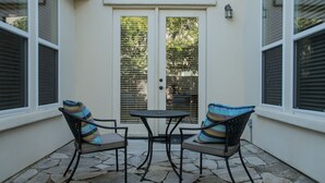 Une terrasse avec deux chaises et une table, une porte blanche et des fenêtres équipées de stores.