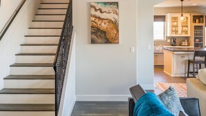 A staircase with a black railing leading to a living area with a sofa and a coffee table. A painting is on the wall, and a kitchen is visible in the background.