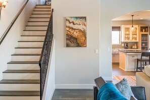A staircase with a black railing leading to a living area with a sofa and a coffee table. A painting is on the wall, and a kitchen is visible in the background.