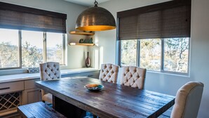 A dining area with a wooden table, four upholstered chairs, a hanging pendant light, and large windows with blinds.