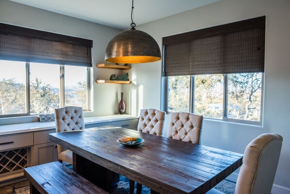 A dining area with a wooden table, four upholstered chairs, a hanging pendant light, and large windows with blinds.