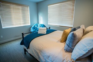 Ein Schlafzimmer mit einem Bett, zwei Fenstern mit Jalousien, einem blauen Stuhl und einem Stapel Handtücher.