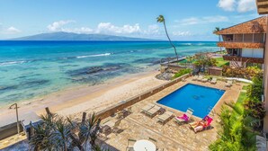 A beachfront property with a swimming pool, lounge chairs, and a view of the ocean and a mountain in the distance.