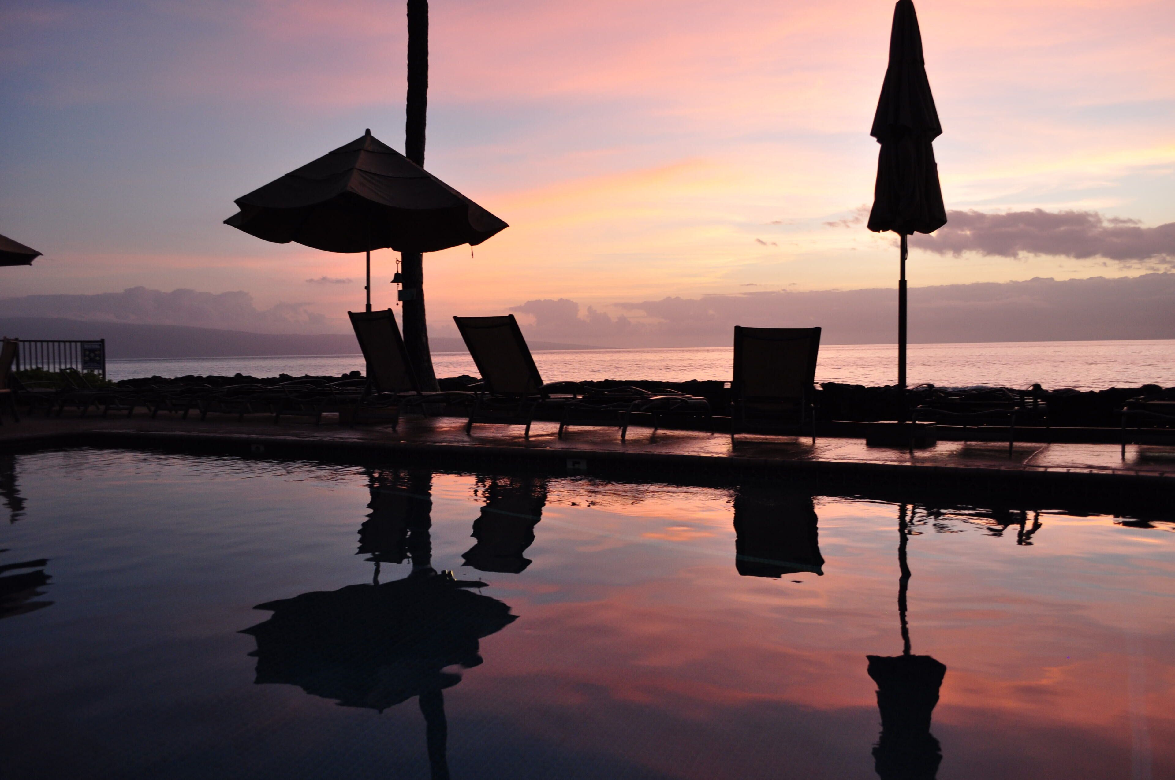 Zona junto a la piscina con sillas de descanso y sombrillas, reflejando una puesta de sol sobre el océano.