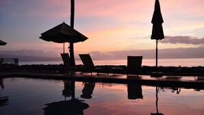 Un espace aménagé au bord de la piscine, avec des chaises longues et des parasols, où se reflète un coucher de soleil sur l’océan.