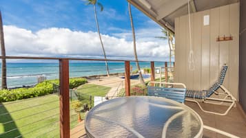 A balcony with a glass table, a blue chair, and a view of the ocean and palm trees.