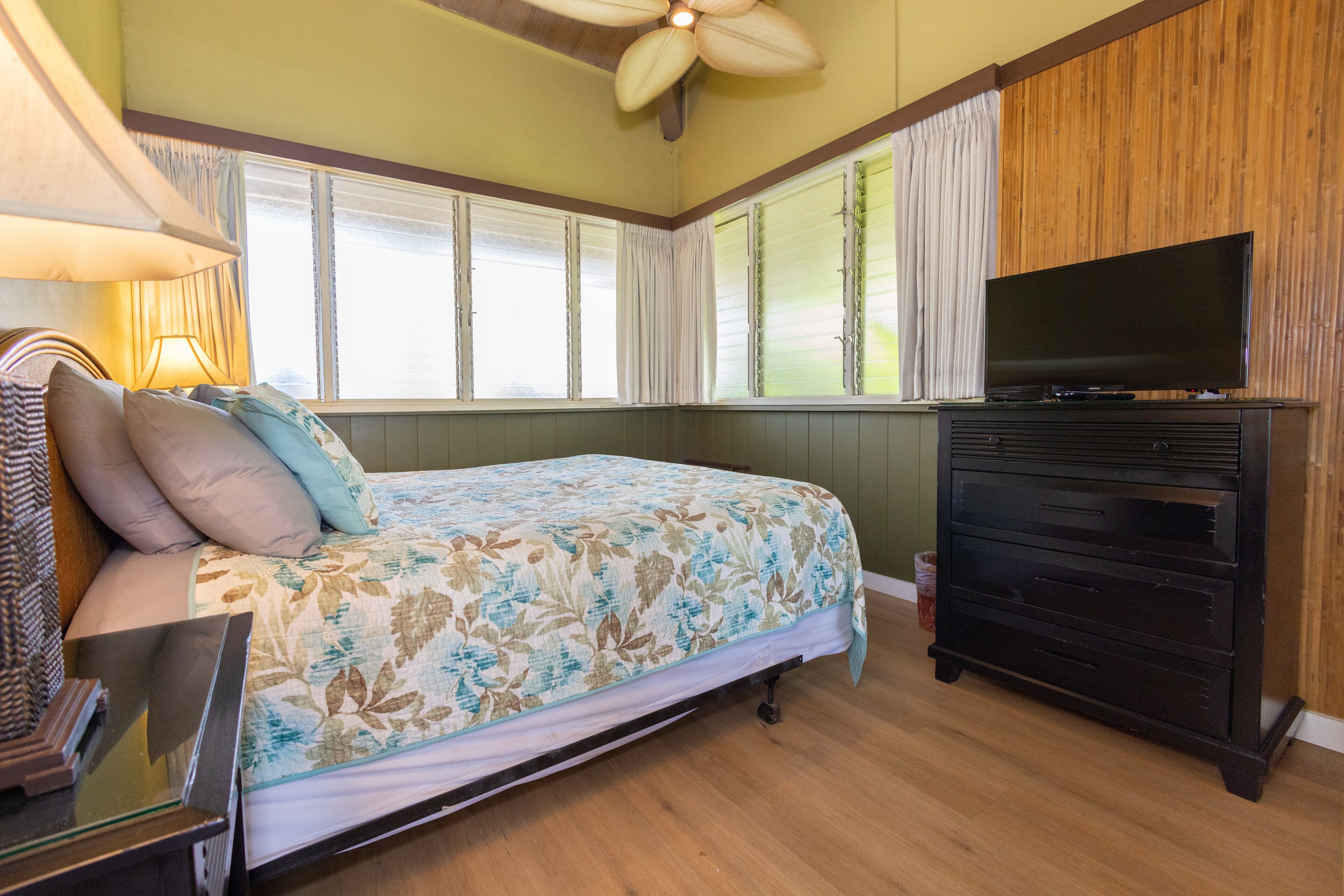 Une chambre à coucher avec un lit, un téléviseur posé sur un meuble, un ventilateur de plafond et une fenêtre avec des rideaux.