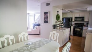 Un coin repas avec une table dressée pour deux personnes, une cuisine avec des meubles blancs et des appareils modernes, et un salon avec un canapé et une guirlande décorative accrochée au mur.