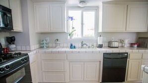 Une cuisine avec des meubles blancs, une cuisinière noire, un lave-vaisselle et une fenêtre agrémentée d’une plante.