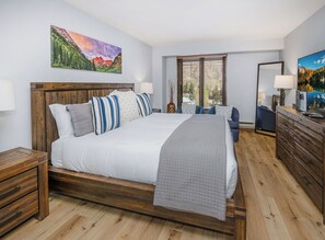 A bedroom with a large bed, wooden headboard, a nightstand, a dresser, and a window with blinds.