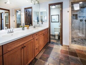 A bathroom with a large wooden vanity, a double sink, a shower area, and a toilet visible through an open door.