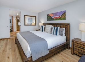 Une chambre à coucher avec un lit en bois, une table de chevet, un tableau accroché au mur et une salle de bain visible à travers une porte ouverte.