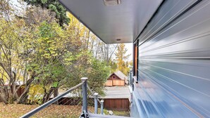 A covered outdoor area with a metal railing, a wooden bench, and a view of trees and a small wooden structure. - Downtown Anchorage Home, 1 Block to Coastal Trail! (Anchorage)