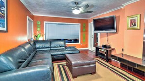 A living room with a sectional sofa, a ceiling fan, a flat-screen TV, and a large window with blinds. - Downtown Anchorage Home, 1 Block to Coastal Trail! (Anchorage)