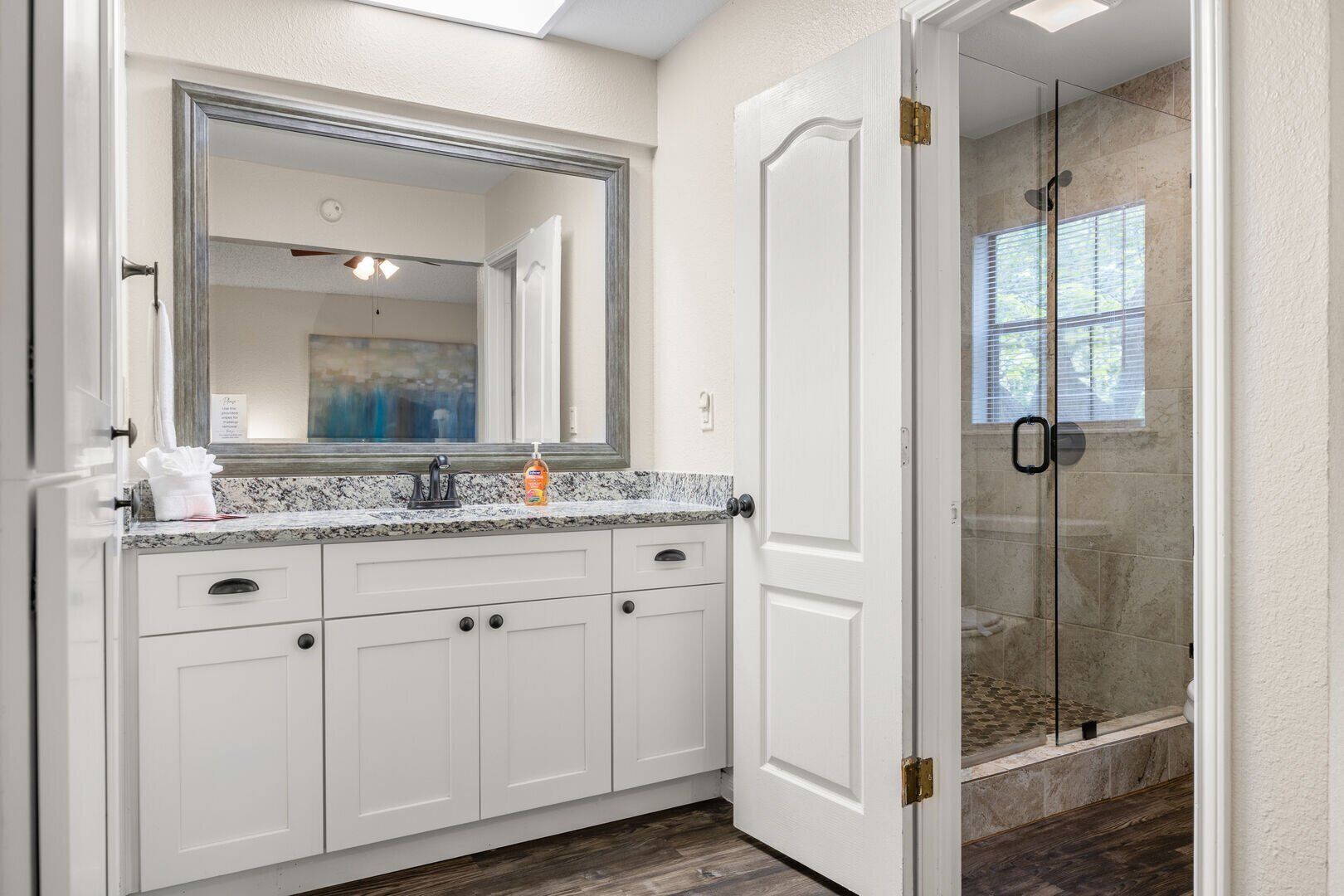 Une salle de bain avec un meuble-lavabo blanc, un grand miroir, une douche avec des portes en verre et une fenêtre laissant entrer la lumière naturelle.