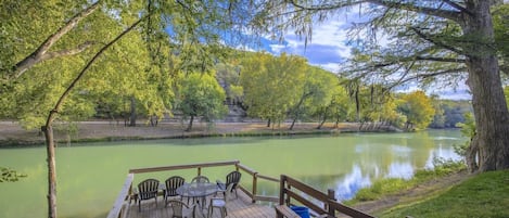 A wooden deck with chairs overlooking a calm river, surrounded by trees and a clear sky.