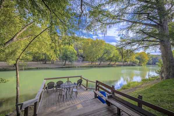 Una terraza de madera con sillas que da a un río tranquilo, rodeada de árboles y un cielo despejado.