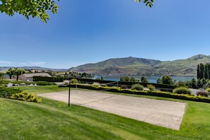 A tennis court with a net, surrounded by well-maintained grass and hedges, overlooking a lake and hills.