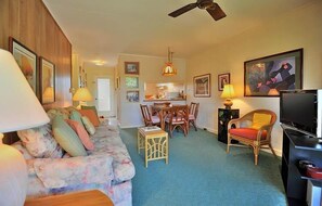 A living room with a sofa, a coffee table, a dining table with chairs, a TV, and a ceiling fan.