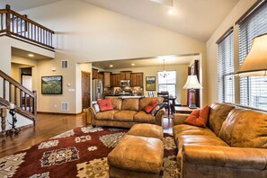 A spacious living area with a leather sofa, ottoman, and a patterned rug. A staircase leads to an upper level, and a kitchen is visible in the background. - Patio, Fire Pit, & Ski Mountain View: Granby Home (Granby)