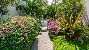 Un sentier bordé d’une végétation luxuriante et de fleurs aux couleurs vives, qui mène à un bâtiment à la façade blanche.