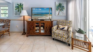 A living room with a television, a patterned armchair, a wooden cabinet, and two framed tropical paintings.