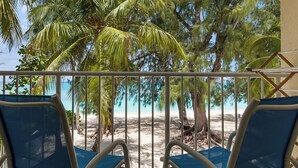 A balcony with two blue chairs, a view of a beach, and palm trees.