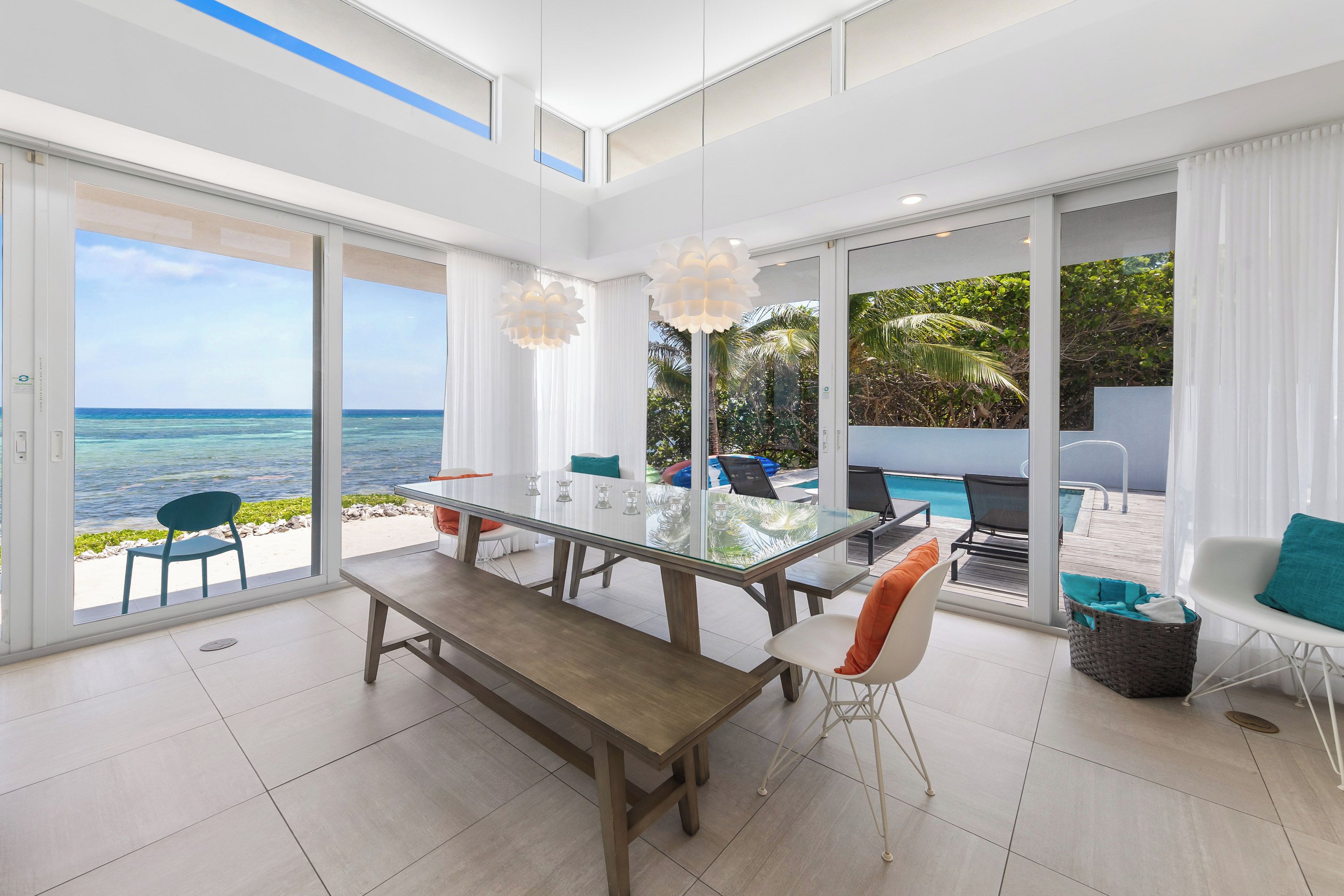 A modern dining area with a glass table, benches, and chairs, overlooking a pool and ocean view.