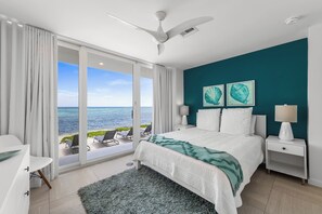 Een slaapkamer met een bed, een nachtkastje, een lamp, een plafondventilator en uitzicht op de oceaan door grote schuifdeuren.