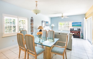 A dining area with a glass table and chairs, a ceiling fan, and a view of the ocean through large windows. - Regal Beach Club 421 - Best Location on 7 Mile Beach! Walk to everything! (Seven Mile Beach)