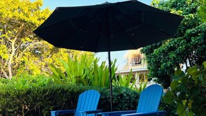 Zwei blaue Stühle und ein schwarzer Sonnenschirm auf sandigem Untergrund, im Hintergrund Grünpflanzen.