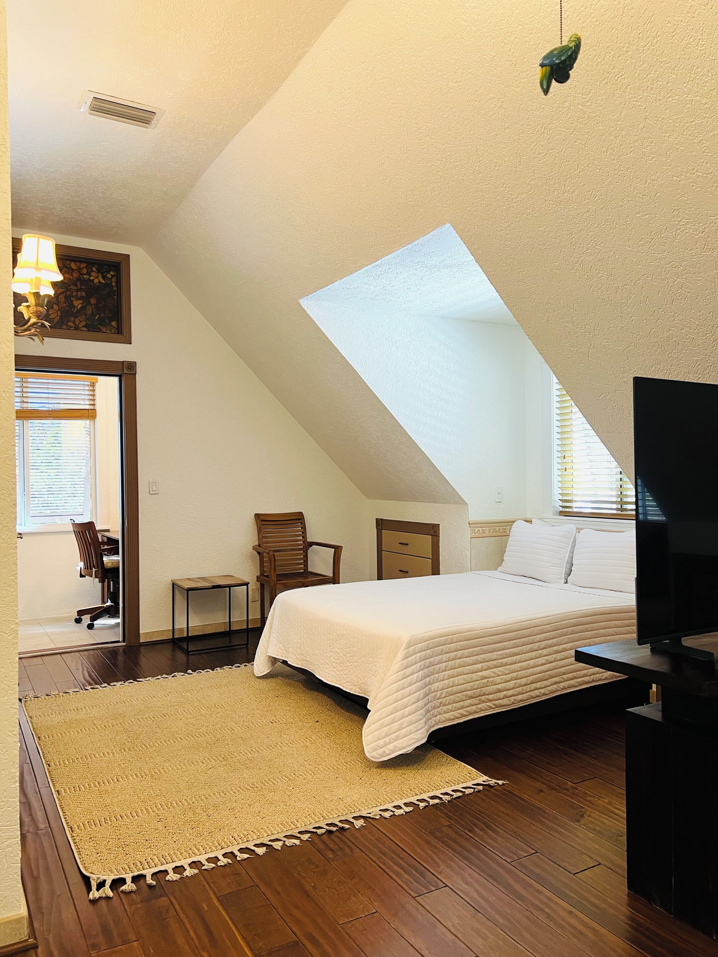 A bedroom with a sloped ceiling, a bed, a desk, a chair, a TV, and a window with blinds.