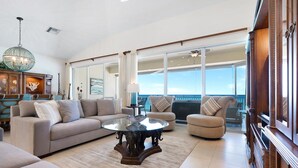 A spacious living room with a large window, a round glass coffee table, and a balcony with a view of the ocean. - Laguna Del Mar 24 -  3rd floor luxury condo on 7MB - Call Cayman Villas! (Seven Mile Beach)