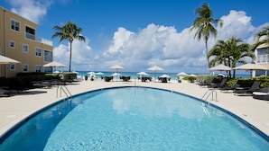 A swimming pool with lounge chairs and umbrellas, surrounded by palm trees and a building with balconies. - Regal Beach Club 623 - Oceanfront Condo with Great Views of the Caribbean Sea (Seven Mile Beach)