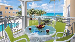 A balcony with a glass table, green chairs, a bowl of fruit, and a magazine. - Regal Beach Club 623 - Oceanfront Condo with Great Views of the Caribbean Sea (Seven Mile Beach)